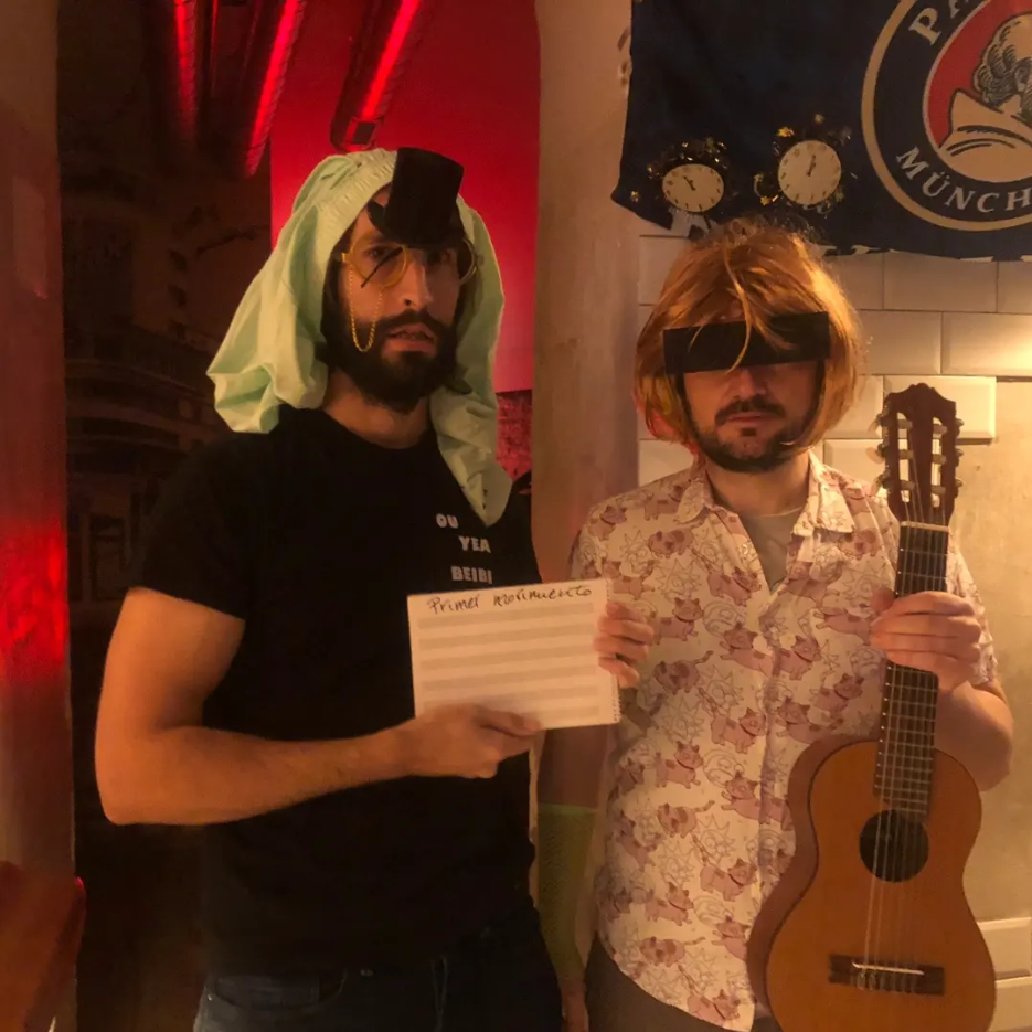 La Mugre: dos integrantes con peluca, gafas de sol y guitarra sosteniendo un cuaderno que dice Primer Movimiento